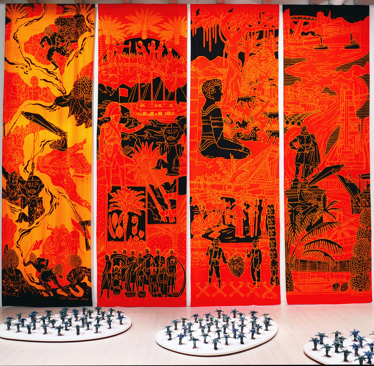 Foto. Fyra höga, avlånga textiler med batiktryck sitter uppsatta bredvid varandra. Trycken är i röd, orange och svart. På golvet framför står tre grupper av många små keramiska skulpturer föreställande palmer. 