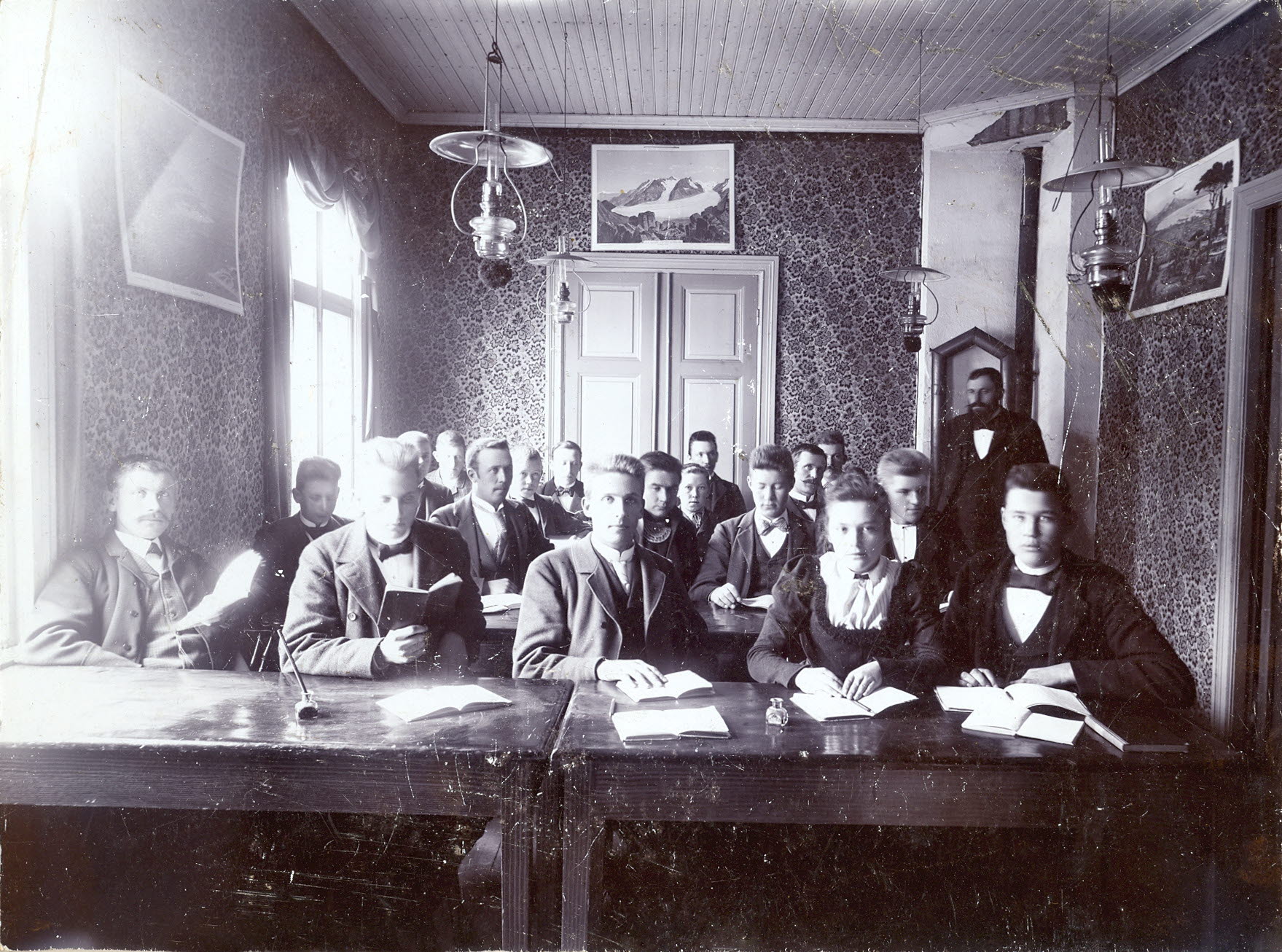 Studerande elever i en gammeldags lektionssal med kakelugn och en fotogenlampa i taket.