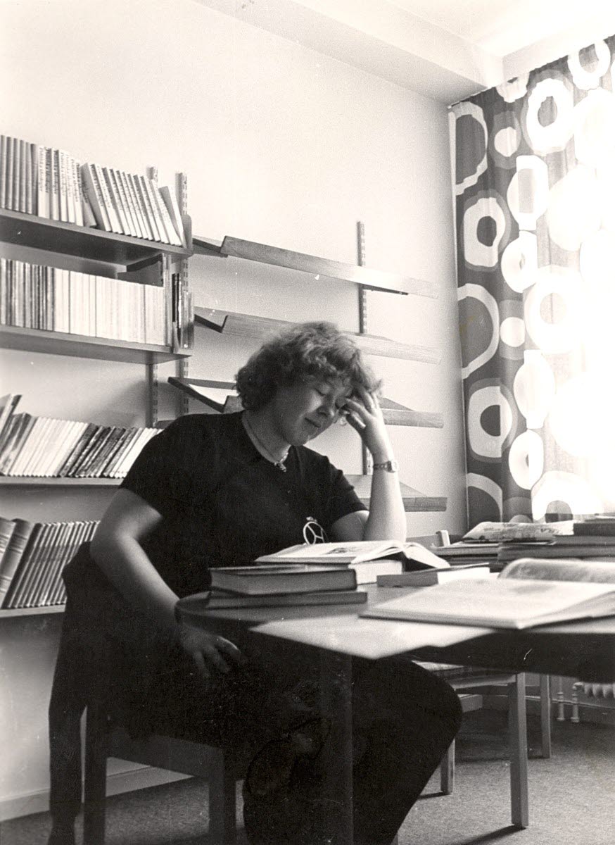 Kvinna med lockig 80-talsfrisyr som sitter i ett rum med bokhyllor och läser i en bok.