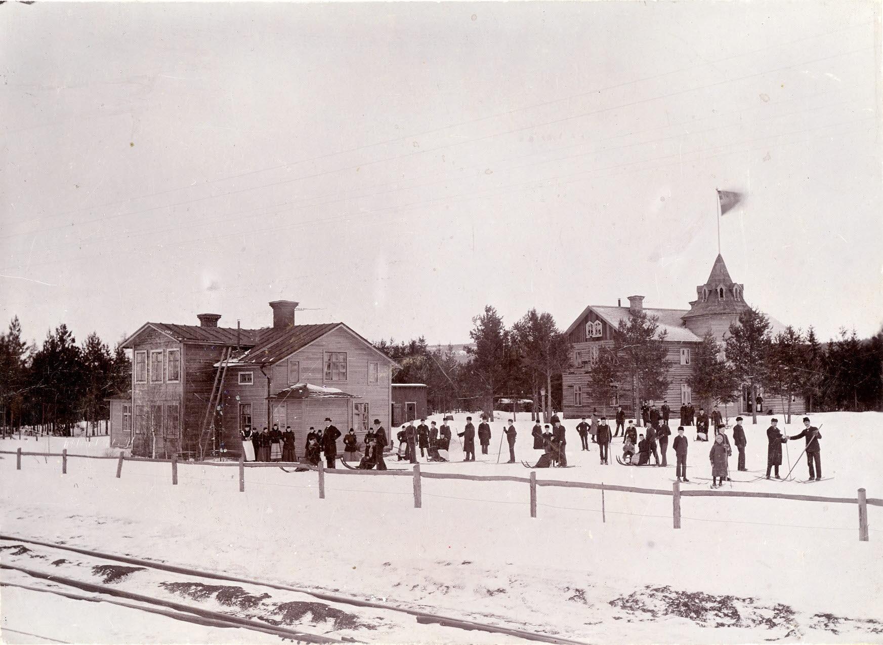 Personer, några på skidor, framför skolbyggnader i trä.