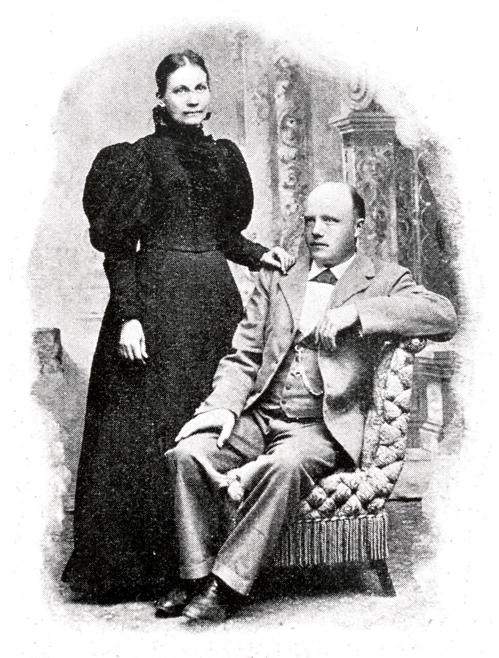 Skallig sittande man i ljus kostym och en stående kvinna i mörk klänning som håller mannen på axeln.