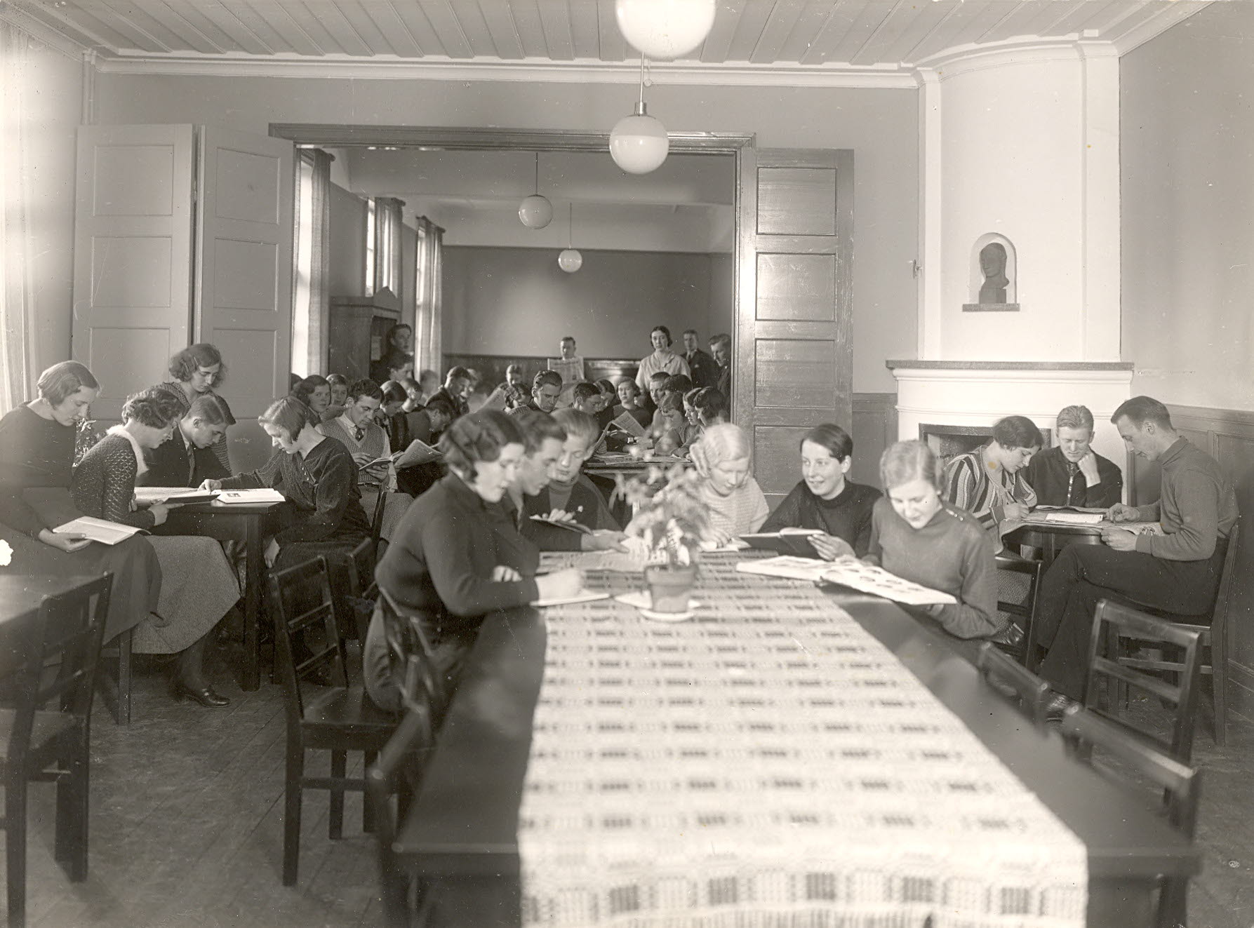 Studerande elever vid långa träbord i ett rum med kakelugn och vita runda glaslampor i taket.