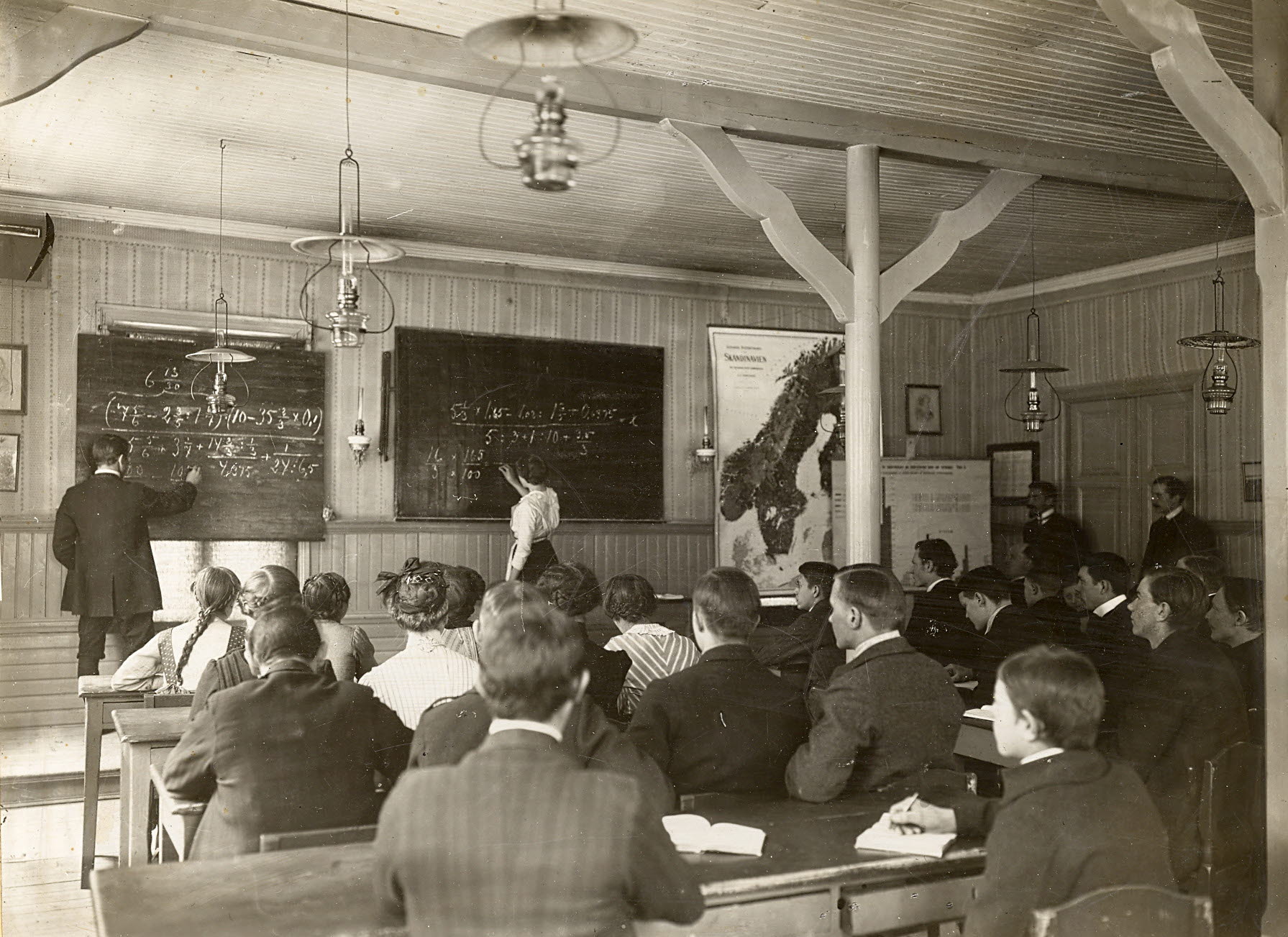 Elever och en undervisande lärare i en lektionssal med två griffeltavlor och några fotogenlampor i taket. 