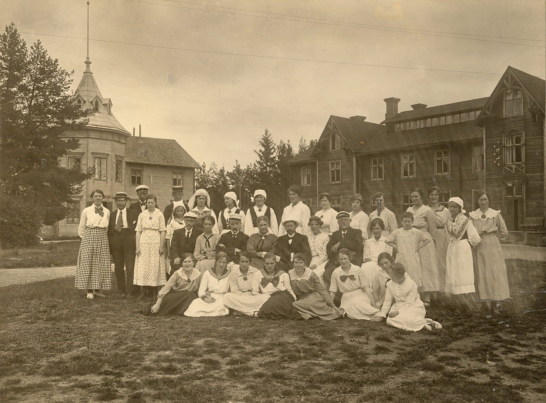 Personer klädda i gammeldags kläder i sommartid samlade för en gruppfotografering framför en träbyggnad. 