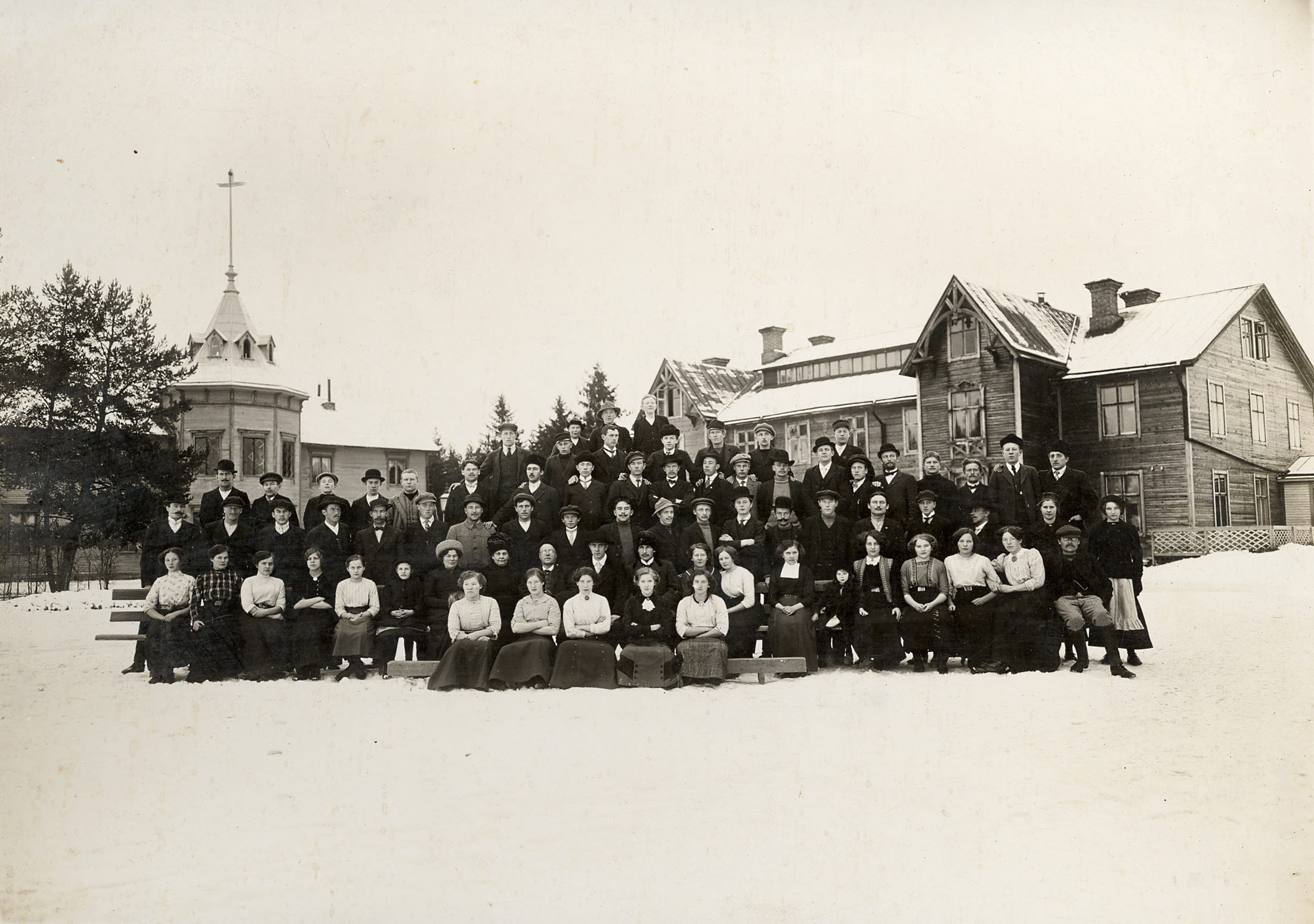 Elever i gammeldags kläder framför två träbyggnader i vintertid.