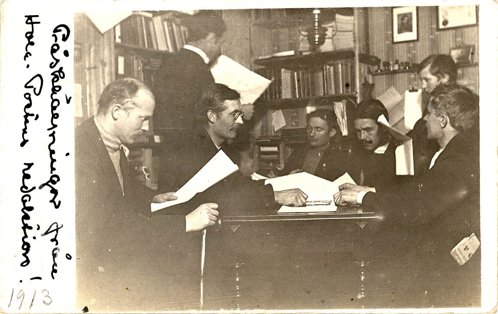 Personer i mörka kostymer som läser tidningar i ett rum med bokhyllor.