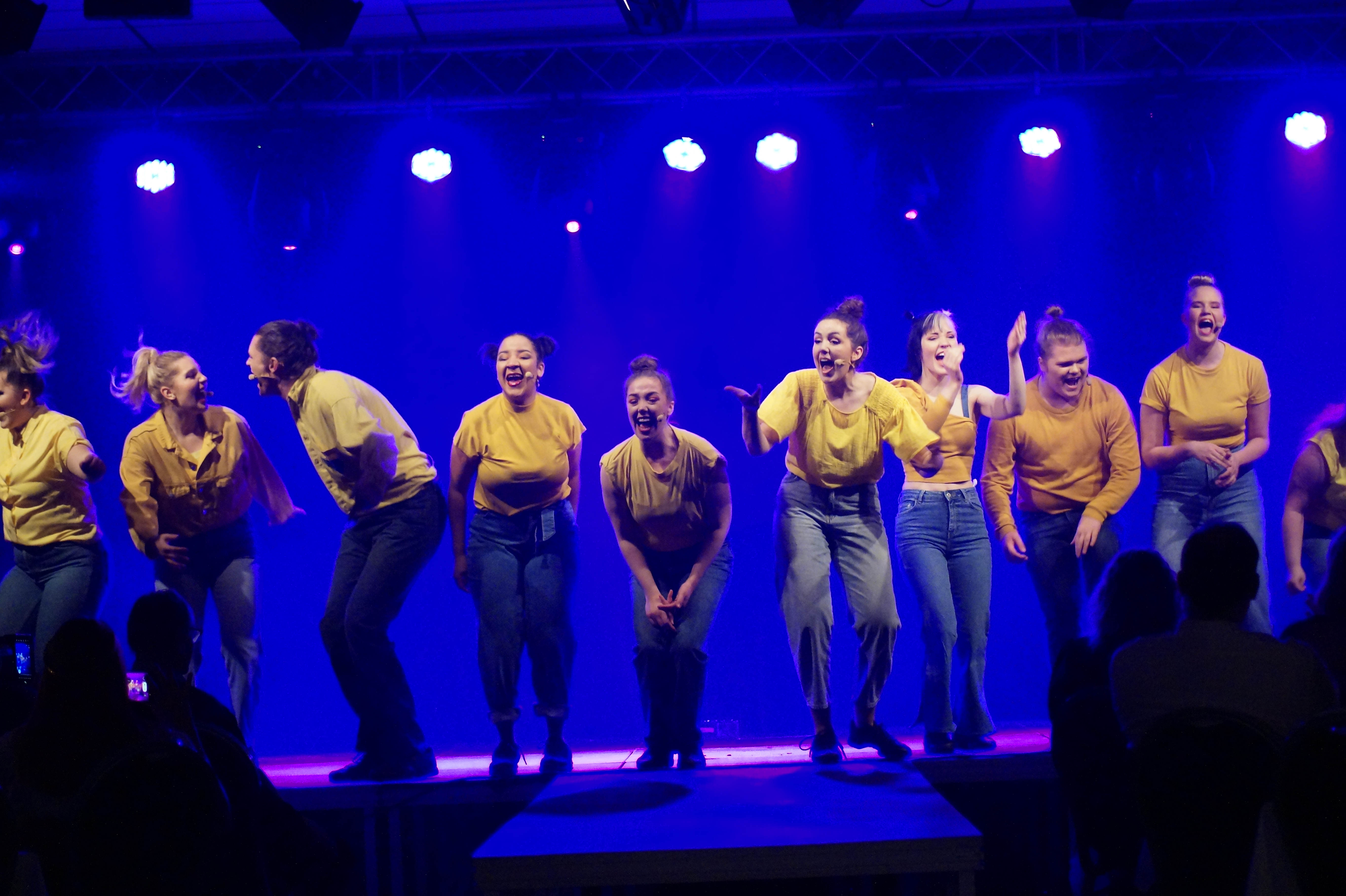 En ensemble klädd i jeans och gula tröjor står på en scen och sjunger. 